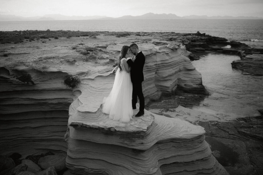 After Wedding Shooting auf Mallorca. Brautpaar steht auf Sandsteinfelsen vor dem Meer und nimmt sich in den Arm.